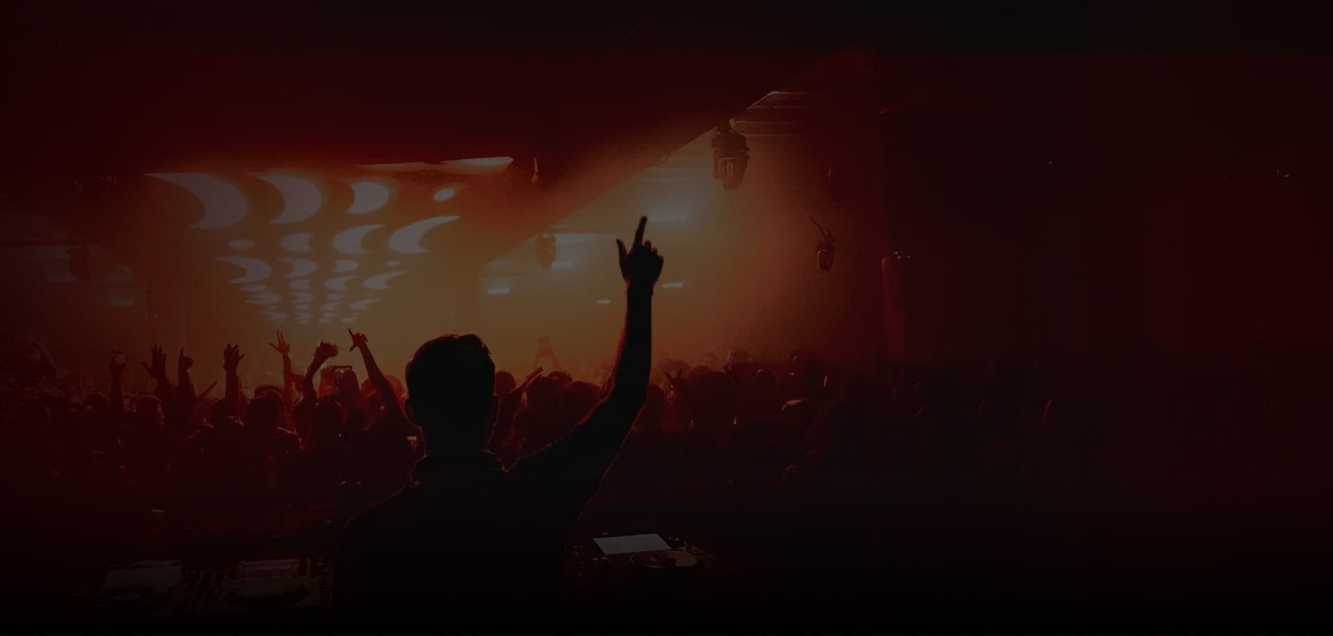 A DJ in silhouette, arm raised over the crowd, under warm red light on the club floor.