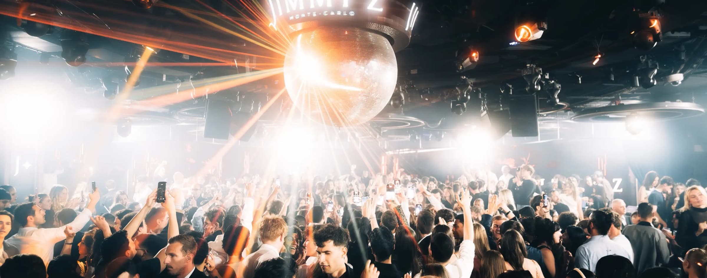 The Jimmy'z Monte-Carlo dancefloor · a packed crowd under a giant mirror ball, light beams and the glowing sign
