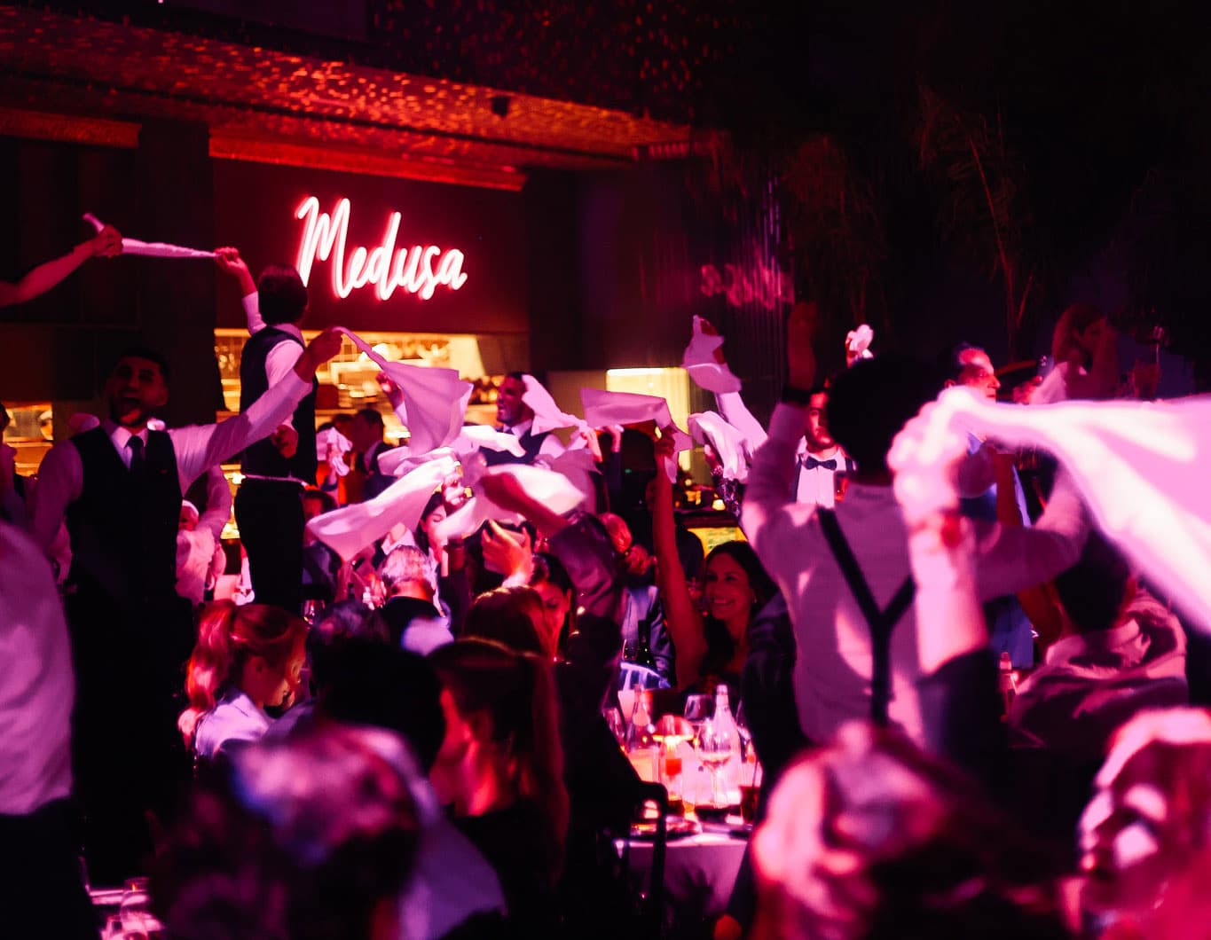 The neon Medusa sign above a full room, waiters in waistcoats waving white napkins under pink light.
