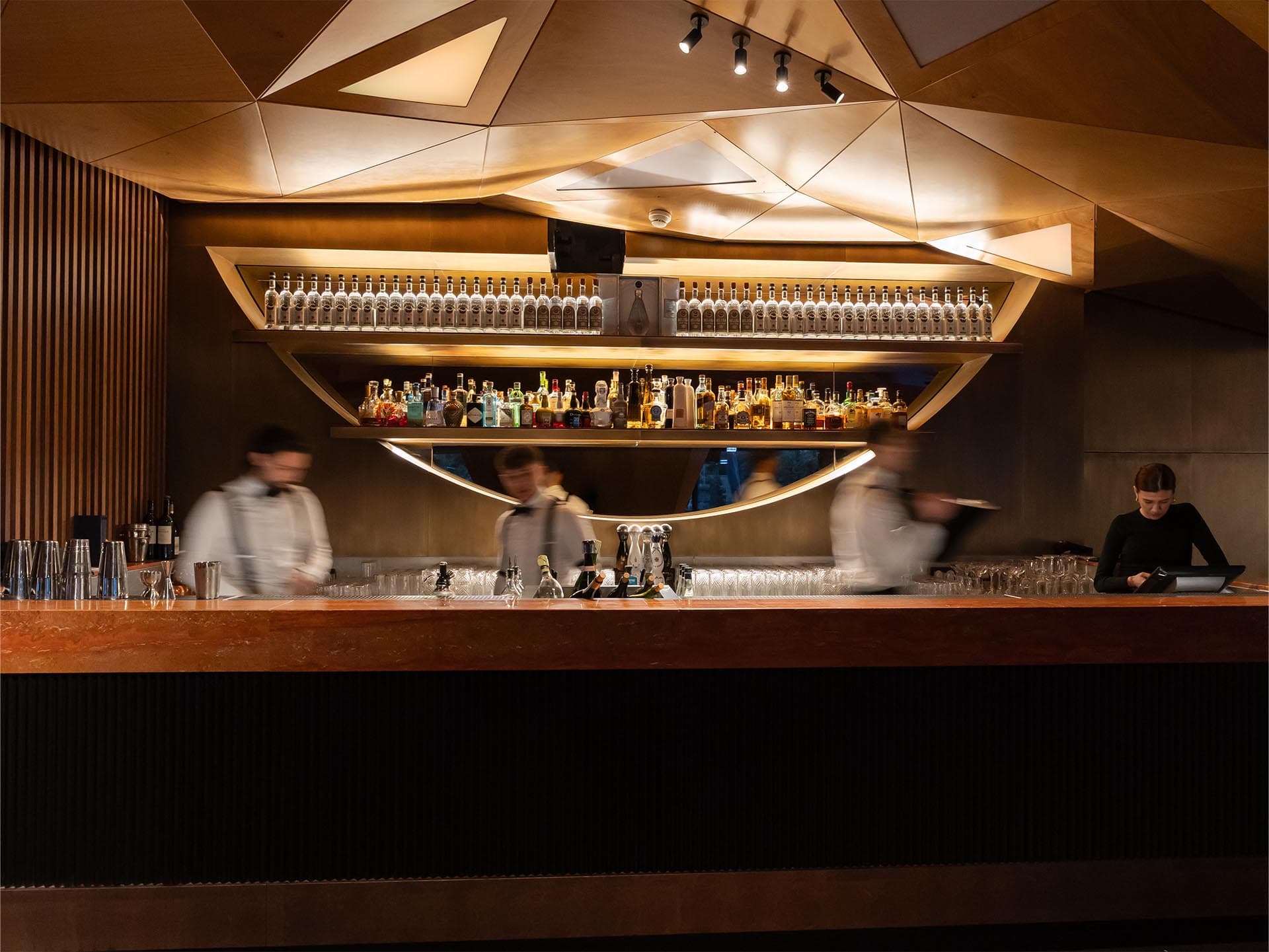 The Twiga Monte-Carlo bar · a faceted timber ceiling above a long backlit counter lined with bottles